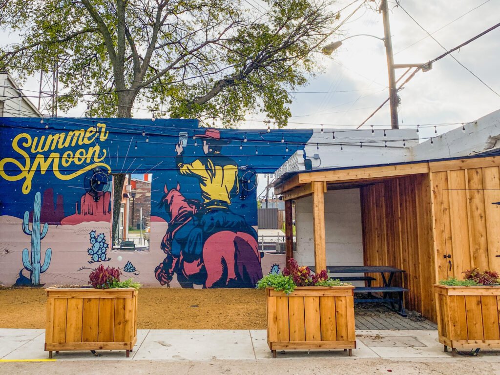 Exterior shot of the seating area behind Summer Moon, a coffee shop in Celina, Texas, featuring a colorful mural painted on the brick wall along one side. 